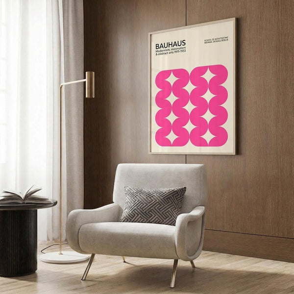May include: Modern living room with beige armchair and geometric pillow, brass floor lamp, dark round side table with open book, and wood-paneled wall displaying framed Bauhaus poster featuring bright pink wavy geometric pattern on cream background.