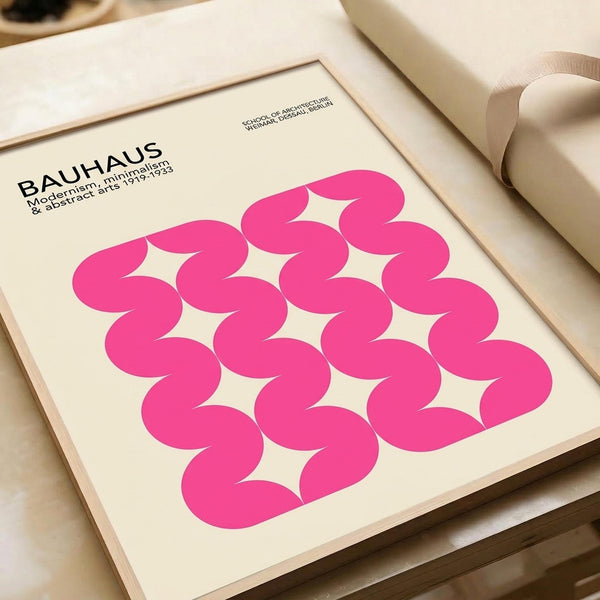 May include: A framed Bauhaus-style poster with bright pink wavy geometric pattern on cream background, displayed on white shelving next to rolled papers and neutral decor. The minimalist print features repeating curved shapes creating an optical wave effect, with black text identifying the German design school and dates 1919-1933.