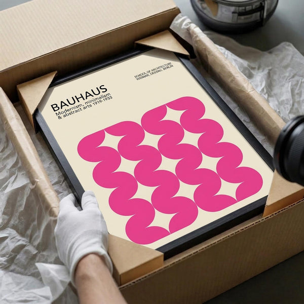 May include: Hands in white gloves carefully unpacking a framed Bauhaus poster from cardboard box with tissue paper. The cream-colored poster features bold pink wavy geometric shapes in a repeating pattern. Black frame sits in brown shipping box on white surface with tape dispenser visible in background.