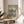 May include: Person holding framed Bauhaus poster with geometric pattern of interlocking curved lines in dark green on cream background. Natural wood frame sits on round wooden side table next to beige upholstered chair. Soft natural lighting creates shadows on neutral wall, with plant leaves visible in corner of minimalist room setting.