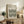 May include: Framed Bauhaus poster with black wavy geometric lines on cream background, displayed on rustic wooden sideboard beside potted fern, ceramic vase, and stacked books in warm-toned living room setting with beige armchair and throw pillows
