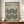 May include: Framed poster with beige background featuring black concentric circle pattern in Bauhaus style, displayed on wooden sideboard next to white ceramic vase and books, with hands holding the brown wooden frame against white paneled wall