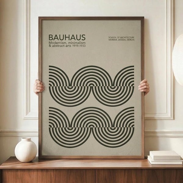 May include: Framed poster with beige background featuring black concentric circle pattern in Bauhaus style, displayed on wooden sideboard next to white ceramic vase and books, with hands holding the brown wooden frame against white paneled wall