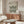 May include: Minimalist Bauhaus poster with geometric wave pattern in dark green on cream background, displayed above wooden credenza in modern living room. White curved chair and glass coffee table visible. Natural lighting creates shadows across walnut furniture and light walls.
