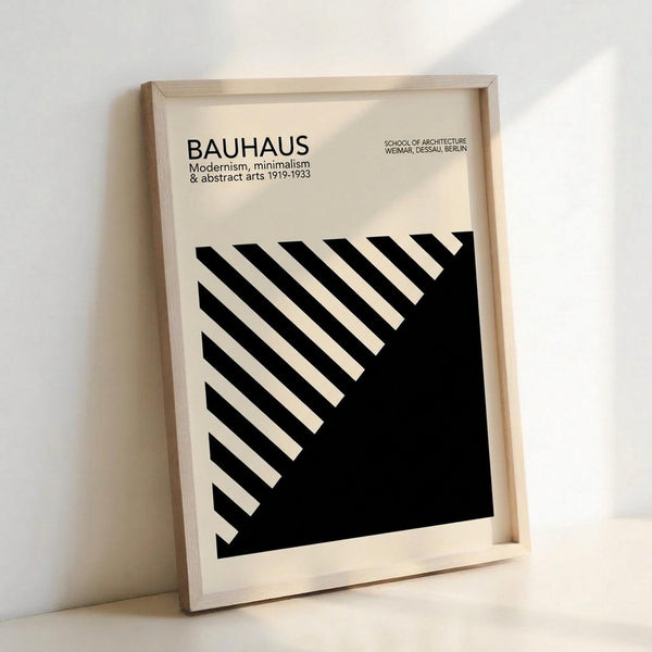 May include: Framed minimalist black and white geometric poster leaning against white wall on light wooden floor. Design features diagonal black stripes creating angular pattern against cream background, with "BAUHAUS" and "Modernism, minimalism & abstract arts 1919-1933" text at top. Natural wood frame casts soft shadows in bright lighting.