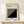 May include: Framed black and white geometric poster with diagonal striped pattern leaning against cream wall on light wood surface, small wrapped gift with twine and greenery sprig positioned in front, minimalist modern interior styling with natural wood frame