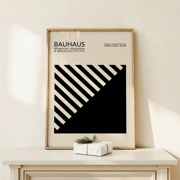May include: Framed black and white geometric poster with diagonal striped pattern leaning against cream wall on light wood surface, small wrapped gift with twine and greenery sprig positioned in front, minimalist modern interior styling with natural wood frame