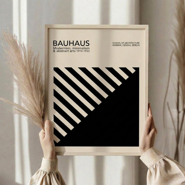 May include: Hands holding a framed minimalist poster with black and white diagonal stripes forming geometric patterns. The print features "BAUHAUS" text and "Modernism, minimalism & abstract arts 1919-1933" with "School of Architecture Weimar, Dessau, Berlin" in smaller text. Light wood frame against cream curtains with dried pampas grass visible on the left side.