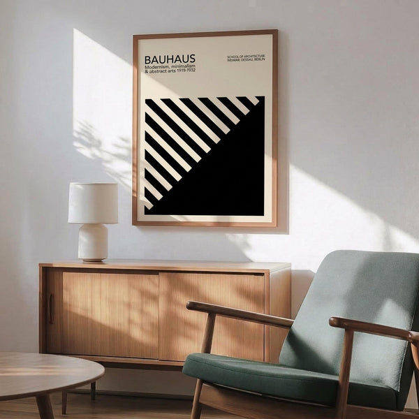 May include: Bauhaus modernist poster with black and white diagonal striped geometric design in wooden frame, hanging above mid-century modern walnut credenza with table lamp, next to sage green upholstered armchair with wooden arms in bright sunlit room with white walls and hardwood floors.