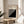 May include: A framed Bauhaus poster with black and white geometric diagonal stripes on cream background, displayed on a round wooden side table next to a beige upholstered chair. Soft natural lighting creates window shadows across the minimalist white wall. Green plant leaves are partially visible in the corner, creating a modern Scandinavian-style interior setting.