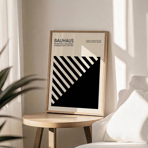 May include: A framed Bauhaus poster with black and white geometric diagonal stripes on cream background, displayed on a round wooden side table next to a beige upholstered chair. Soft natural lighting creates window shadows across the minimalist white wall. Green plant leaves are partially visible in the corner, creating a modern Scandinavian-style interior setting.