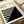 May include: Minimalist black and white geometric poster with angular striped pattern leaning against beige upholstered chair. Clean modern design features bold triangular shapes and linear elements in monochrome palette. Artwork displayed in simple light wood frame on neutral background with rolled papers nearby.