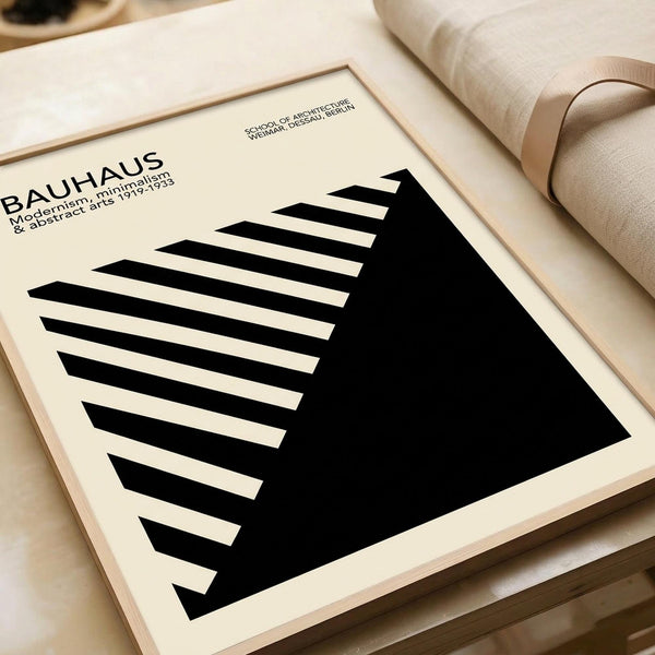 May include: Minimalist black and white geometric poster with angular striped pattern leaning against beige upholstered chair. Clean modern design features bold triangular shapes and linear elements in monochrome palette. Artwork displayed in simple light wood frame on neutral background with rolled papers nearby.
