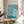 May include: Framed poster with flowing turquoise and cream swirled pattern displayed on light wood round side table next to beige upholstered chair. Bright natural light streams across white walls creating soft shadows, with green plant leaves visible in corner of minimalist modern room setting.