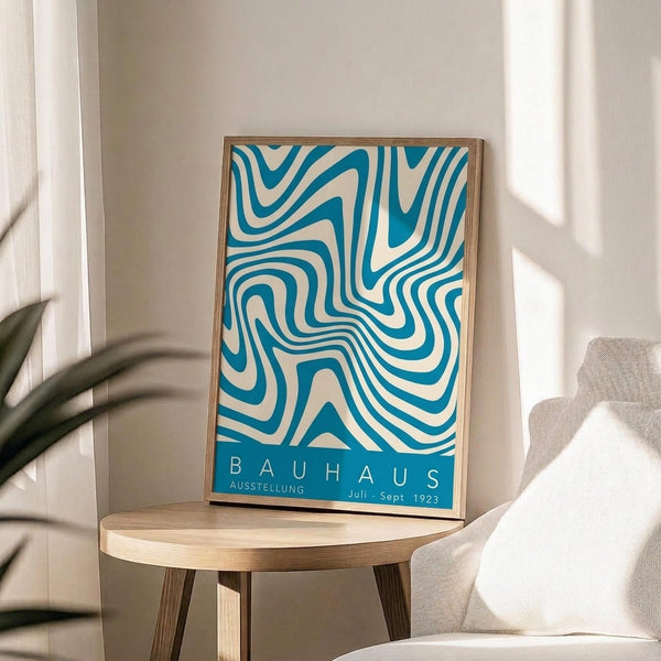 May include: Framed poster with flowing turquoise and cream swirled pattern displayed on light wood round side table next to beige upholstered chair. Bright natural light streams across white walls creating soft shadows, with green plant leaves visible in corner of minimalist modern room setting.
