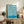 May include: Framed abstract poster with flowing blue and cream curved lines in a wavy pattern, displayed on a rustic wooden sideboard next to a fern plant, ceramic vase, and stacked books in a cozy living room setting with neutral tones and natural lighting.