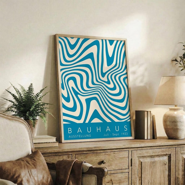 May include: Framed abstract poster with flowing blue and cream curved lines in a wavy pattern, displayed on a rustic wooden sideboard next to a fern plant, ceramic vase, and stacked books in a cozy living room setting with neutral tones and natural lighting.