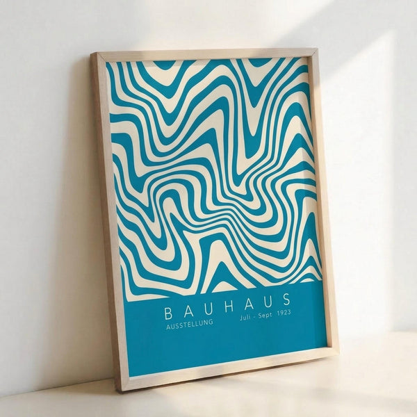 A framed poster with swirling teal and cream abstract patterns creating flowing, wave-like designs. The bottom features a solid teal section with white text reading "BAUHAUS" and "AUSSTELLUNG Juli - Sept 1923". The print is displayed in a light wooden frame leaning against a white wall, showcasing the mid-century modern aesthetic with organic, undulating lines that create an optical illusion effect.
