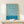 May include: Framed Bauhaus exhibition poster with flowing teal and cream swirled pattern, displayed on light wood surface next to small wrapped gift with twine and dried botanicals, against neutral wall with soft natural lighting
