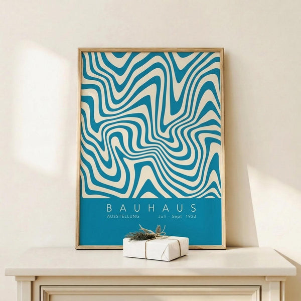 May include: Framed Bauhaus exhibition poster with flowing teal and cream swirled pattern, displayed on light wood surface next to small wrapped gift with twine and dried botanicals, against neutral wall with soft natural lighting