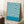 May include: Framed vintage-style Bauhaus exhibition poster with flowing blue and cream swirled pattern, displayed leaning against white wall next to wooden side table with coffee cup, on textured cream rug with beige throw blanket visible in corner
