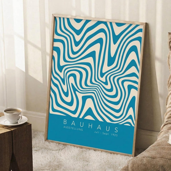 May include: Framed vintage-style Bauhaus exhibition poster with flowing blue and cream swirled pattern, displayed leaning against white wall next to wooden side table with coffee cup, on textured cream rug with beige throw blanket visible in corner