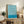May include: Framed Bauhaus exhibition poster with flowing blue and white wavy lines displayed on rustic wooden sideboard alongside fern plant, ceramic vase, and books in warm-toned living room setting with beige armchair