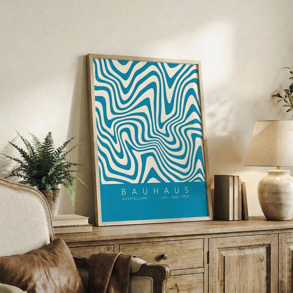 May include: Framed Bauhaus exhibition poster with flowing blue and white wavy lines displayed on rustic wooden sideboard alongside fern plant, ceramic vase, and books in warm-toned living room setting with beige armchair