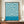 May include: Framed Bauhaus exhibition poster with swirling blue and cream organic patterns displayed on wooden sideboard next to white ceramic vase and books. The minimalist mid-century modern interior features white paneled walls and warm wood furniture. The poster shows flowing, wave-like designs in teal blue against cream background with exhibition text at bottom.