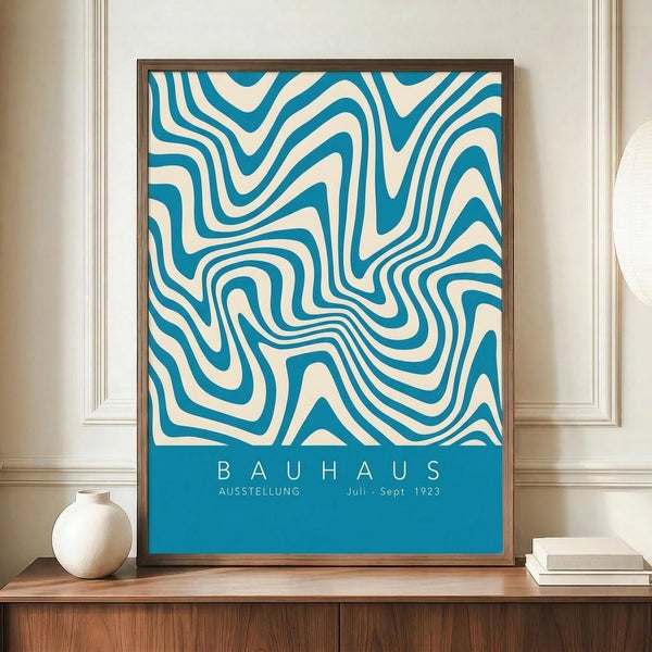 May include: Framed Bauhaus exhibition poster with swirling blue and cream organic patterns displayed on wooden sideboard next to white ceramic vase and books. The minimalist mid-century modern interior features white paneled walls and warm wood furniture. The poster shows flowing, wave-like designs in teal blue against cream background with exhibition text at bottom.