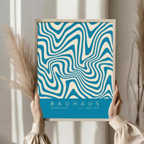 May include: Person holding framed poster with blue and cream wavy optical pattern design in thin light wood frame. Bauhaus exhibition text at bottom in white letters on blue band. Dried pampas grass visible to left, neutral beige clothing and white curtained background create minimalist styling.
