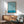 May include: Framed poster with teal and cream wavy geometric pattern displayed above wooden sideboard with table lamp, shown next to sage green mid-century armchair and round coffee table in modern living room with natural sunlight casting shadows on white wall.