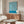 May include: Blue and white swirling abstract pattern poster hanging above a mid-century modern wooden sideboard in a minimalist living room. The artwork features flowing organic lines in teal blue against cream background. Room styled with white curved chair, glass oval coffee table, modern white table lamp, and copper floor lamp on light wooden floors.