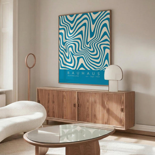 May include: Blue and white swirling abstract pattern poster hanging above a mid-century modern wooden sideboard in a minimalist living room. The artwork features flowing organic lines in teal blue against cream background. Room styled with white curved chair, glass oval coffee table, modern white table lamp, and copper floor lamp on light wooden floors.