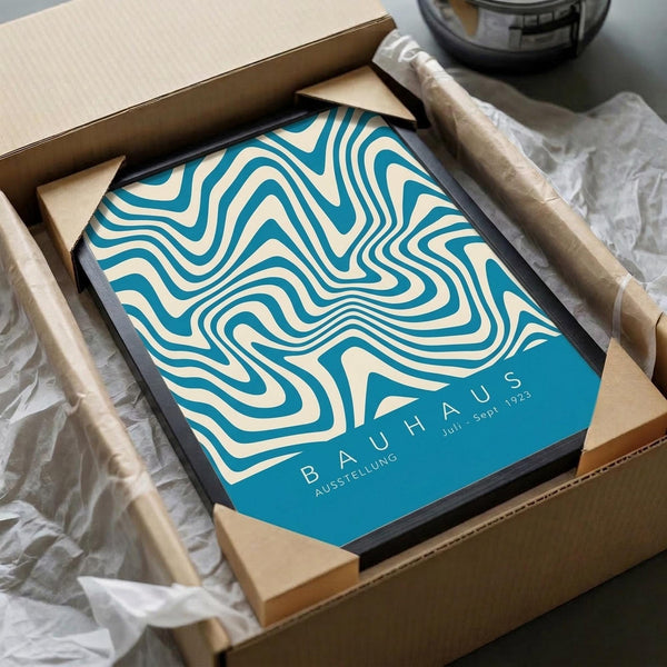 May include: Framed poster with flowing teal and cream wavy lines in an optical illusion pattern, displayed in black frame inside cardboard shipping box with protective packaging. Text reads "BAUHAUS AUSSTELLUNG Juli - Sept. 1923" in bottom corner. Surrounded by white tissue paper and brown packing materials on light surface.