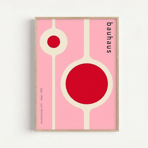 Framed modernist poster with pink background featuring two geometric circular designs with red centers connected by cream-colored stems. Black text reads "bauhaus" vertically on right side and "Ausstellung Juli Sept. 1923" vertically on left. Light wood frame against white wall.