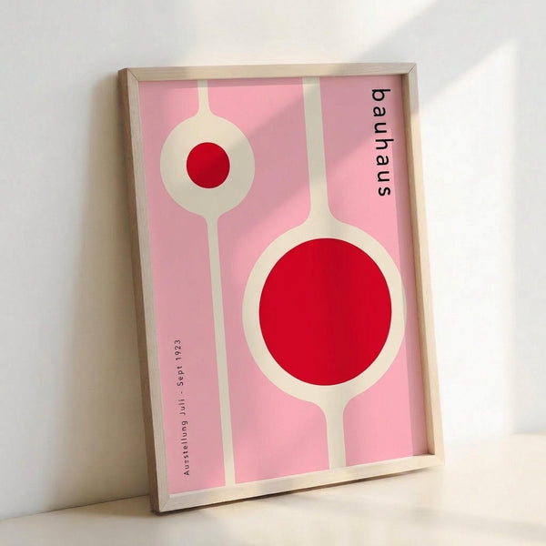 Framed modernist poster with pink background featuring two abstract circular forms in cream and red - one small circle at top, one large circle at bottom, connected by vertical stems. Black text reads "bauhaus" vertically on right side and "Ausstellung Juli - Sept 1923" on left. Light wood frame displayed against white wall.