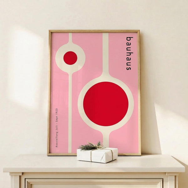 May include: Framed pink poster with geometric red circles and white lines in Bauhaus style, displayed on light wood console table next to small wrapped gift with twine and evergreen sprig against cream wall