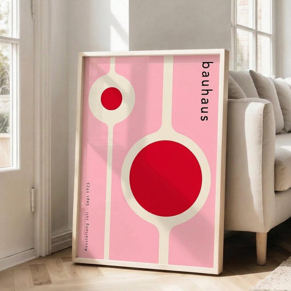 May include: Framed geometric art print with pink background featuring white linear shapes and red circles in Bauhaus style, displayed against white wall next to beige upholstered chair in bright living room with wood floors and large windows