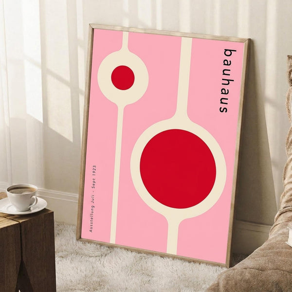 May include: Framed pink poster with geometric white circles containing red centers, displayed in modern room setting. Artwork features Bauhaus-style design with cream/white connecting elements on bright pink background. Wooden frame leans against white wall on textured cream rug, with coffee cup on wooden block nearby and beige fabric visible.