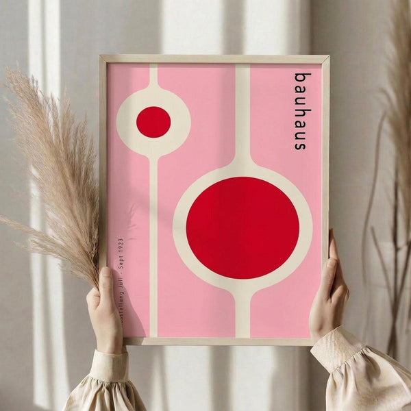 May include: Woman in cream sweater holding framed Bauhaus-style poster with pink background, white geometric circles containing red dots, displayed against white curtained window with dried pampas grass bundle visible to left side.