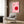 May include: Pink and red geometric Bauhaus-style poster hanging on wood-paneled wall above beige upholstered armchair with patterned pillow. Modern brass floor lamp and dark round side table with open book nearby. Neutral tones with white curtains and wood flooring create contemporary living room setting.