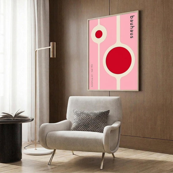 May include: Pink and red geometric Bauhaus-style poster hanging on wood-paneled wall above beige upholstered armchair with patterned pillow. Modern brass floor lamp and dark round side table with open book nearby. Neutral tones with white curtains and wood flooring create contemporary living room setting.