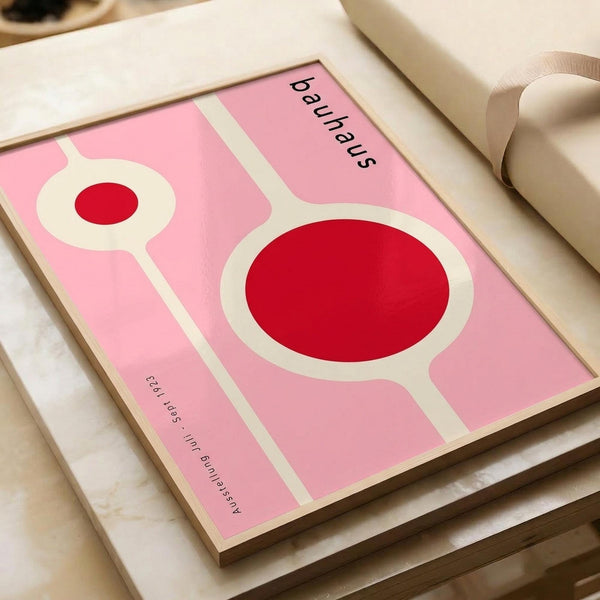May include: Pink and red geometric Bauhaus-style poster print displayed on white surface next to beige ribbon roll. Abstract design features two circular forms with white outlines on pink background, black text reading "bauhaus" and smaller text below. Modern minimalist art print shown in styled flat lay photography setup.