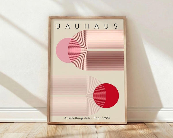 May include: Framed Bauhaus-style poster with geometric design featuring red and pink circular forms intersected by curved parallel lines on cream background, displayed in light wood frame against white wall on wooden floor with natural lighting shadows