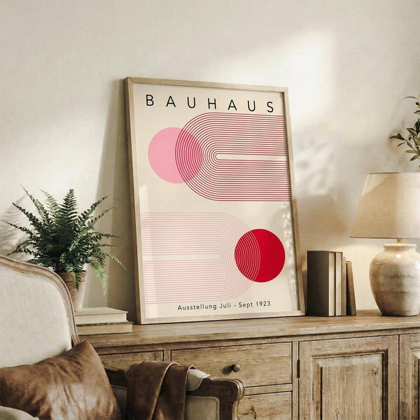 May include: Framed Bauhaus exhibition poster with geometric red and pink shapes and curved lines displayed on rustic wooden sideboard alongside fern plant, ceramic vase, and books in cozy living room setting with leather furniture and neutral wall