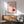 May include: Bauhaus exhibition poster from 1923 featuring geometric design with pink and red circles, curved linear patterns on cream background, displayed in wooden frame above mid-century modern furniture including teal upholstered chair and wooden sideboard with ceramic lamp, natural sunlight casting plant shadows on white wall