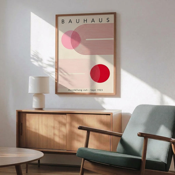 May include: Bauhaus exhibition poster from 1923 featuring geometric design with pink and red circles, curved linear patterns on cream background, displayed in wooden frame above mid-century modern furniture including teal upholstered chair and wooden sideboard with ceramic lamp, natural sunlight casting plant shadows on white wall