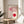 May include: Framed Bauhaus exhibition poster with geometric design featuring pink and red circles with curved line patterns, displayed on wooden round table next to beige upholstered chair. Natural lighting creates soft shadows on neutral wall background with plant leaves visible in corner.