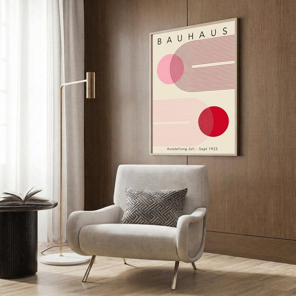 May include: Vintage-style Bauhaus poster with geometric pink and red circular designs and linear patterns hanging on wood wall panel above beige upholstered armchair with patterned pillow. Modern brass floor lamp and dark round side table with open book visible. Warm neutral interior with curtained window and wood flooring.